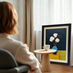 A black framed art print of white flowers in a blue vase, next to a small wooden table with a white teacup, in a sunlit interior with curtains.