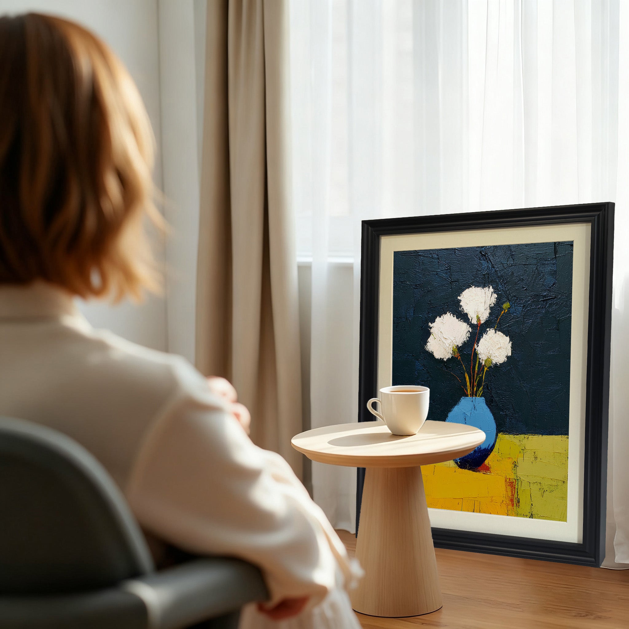 A black framed art print of white flowers in a blue vase, next to a small wooden table with a white teacup, in a sunlit interior with curtains.