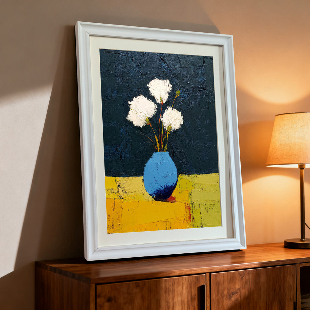 Still life art print featuring white flowers in a bright blue vase against a dark blue background and a yellow table, displayed in a white wood frame on a dark wood cabinet with a lamp on the side.
