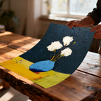 A person's hands holding a rolled, unframed art print of white flowers in a blue vase over a wooden table, showing the textured brushstrokes.