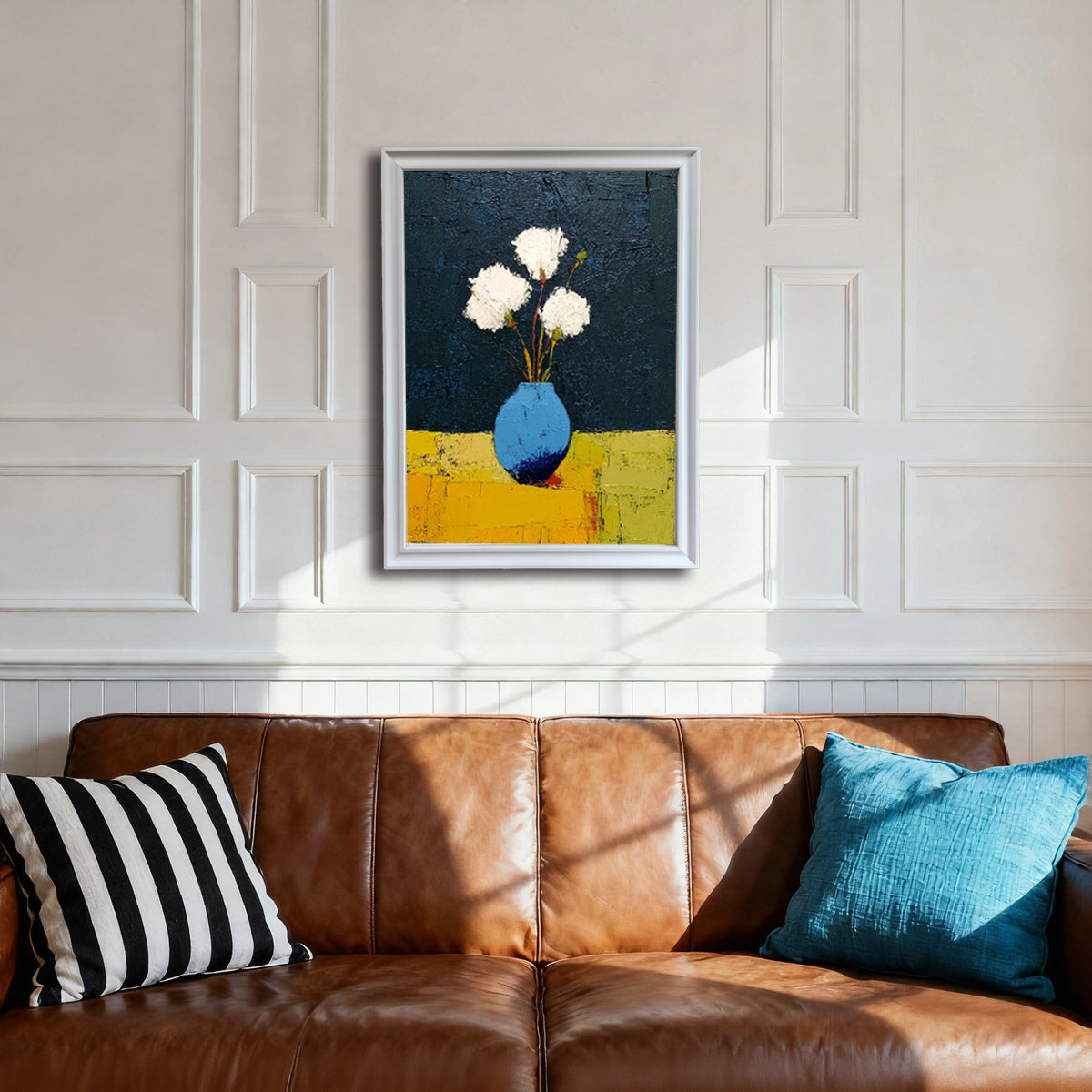 Art print of white flowers in a blue vase in a white wood frame, hanging on a white wainscoting wall above a brown leather sofa with black-and-white and blue pillows.