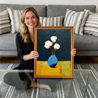Woman sitting on the floor holding a framed art print of white flowers in a blue vase, framed in a gold-colored wood frame. Gray couch with striped pillows in the background.