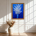 A vertical, gold-framed painting of silver-white flowers in a blue vase against a royal blue background, hanging on a white wainscoted wall. A large, off-white ceramic vase with dried flowers sits on the wooden floor below it, with strong sunlight creating window shadows.
