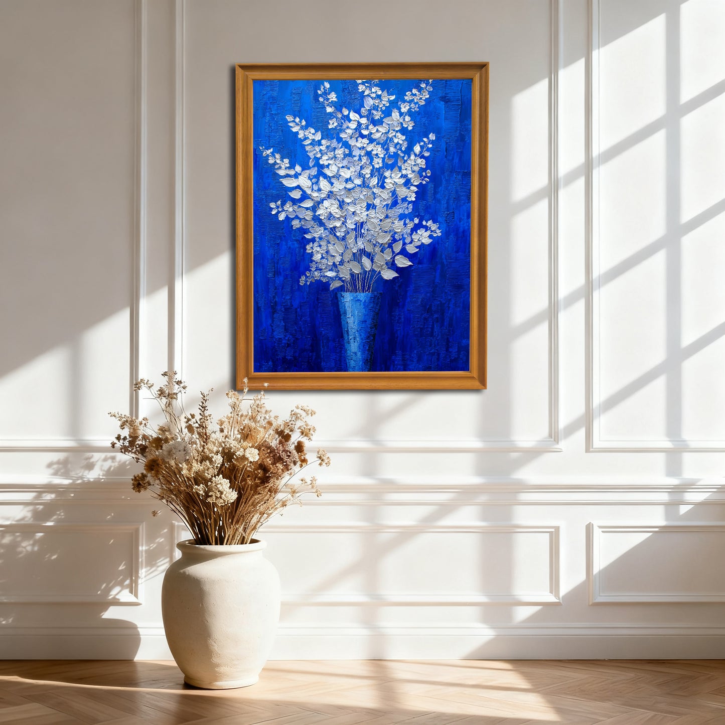 A vertical, gold-framed painting of silver-white flowers in a blue vase against a royal blue background, hanging on a white wainscoted wall. A large, off-white ceramic vase with dried flowers sits on the wooden floor below it, with strong sunlight creating window shadows.