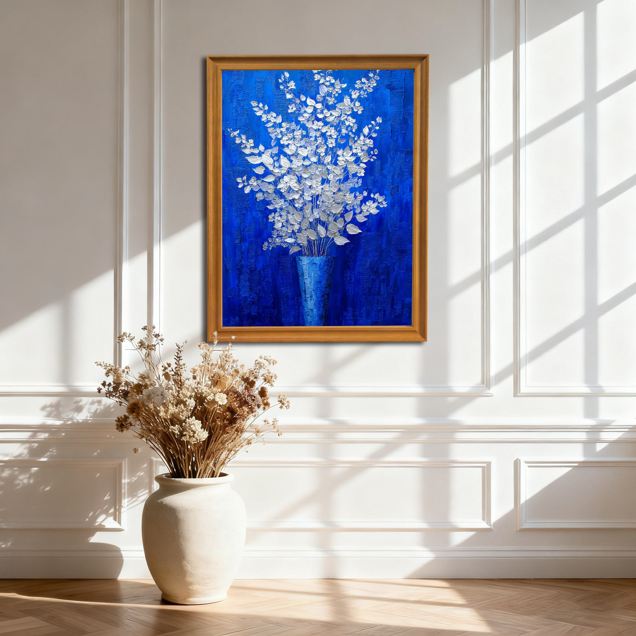 A vertical, gold-framed painting of silver-white flowers in a blue vase against a royal blue background, hanging on a white wainscoted wall. A large, off-white ceramic vase with dried flowers sits on the wooden floor below it, with strong sunlight creating window shadows.