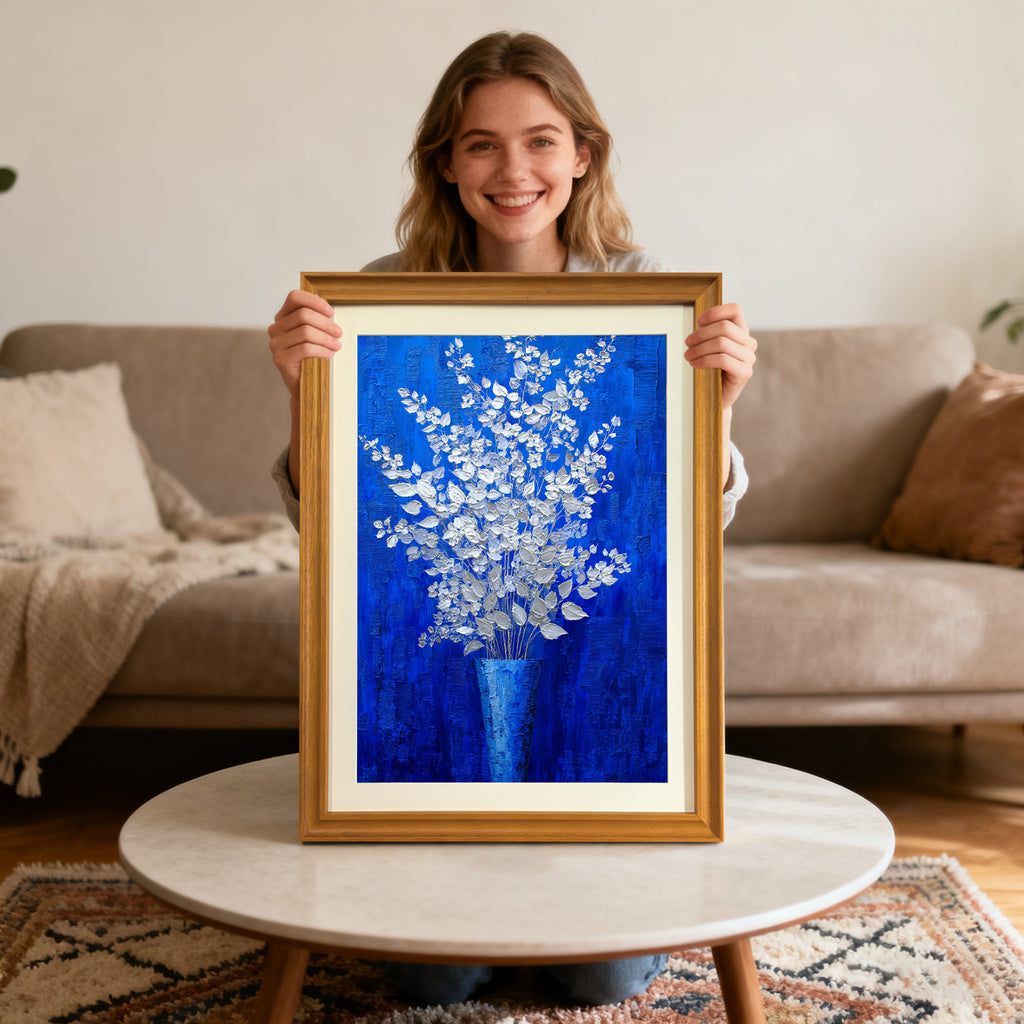 A smiling young woman sitting on the floor behind a coffee table holds up a gold-framed painting of silver-white flowers in a blue vase against a royal blue background.