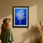 A young woman with red, curly hair and a green headband stands in an earth-toned room looking up at a vertical, black-framed painting of silver-white flowers on a royal blue background hanging on the wall. A tall, woven vase with dried grasses is on the right.