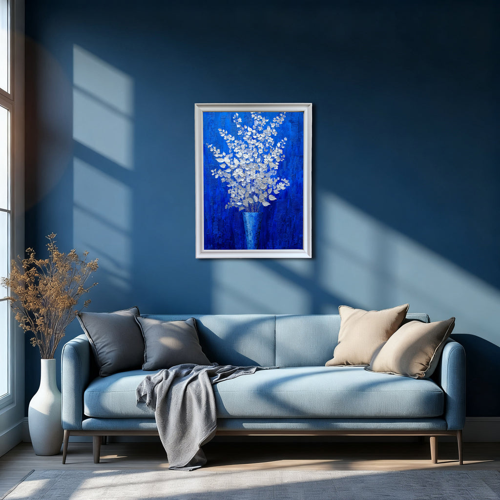 A vertical, white-framed painting of silver-white flowers on a royal blue background hanging above a light blue sofa with beige and gray pillows, in a modern room with dark blue walls and large window shadows.
