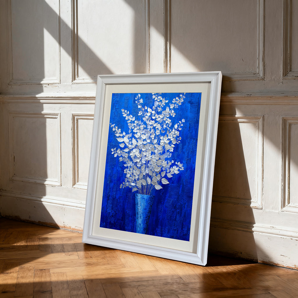 A vertical, framed oil painting of silver and white flowers in a blue vase against a textured royal blue background. The painting is in a thick white frame and is leaning against a white wainscoted wall on a hardwood floor, with strong sunlight casting shadows.