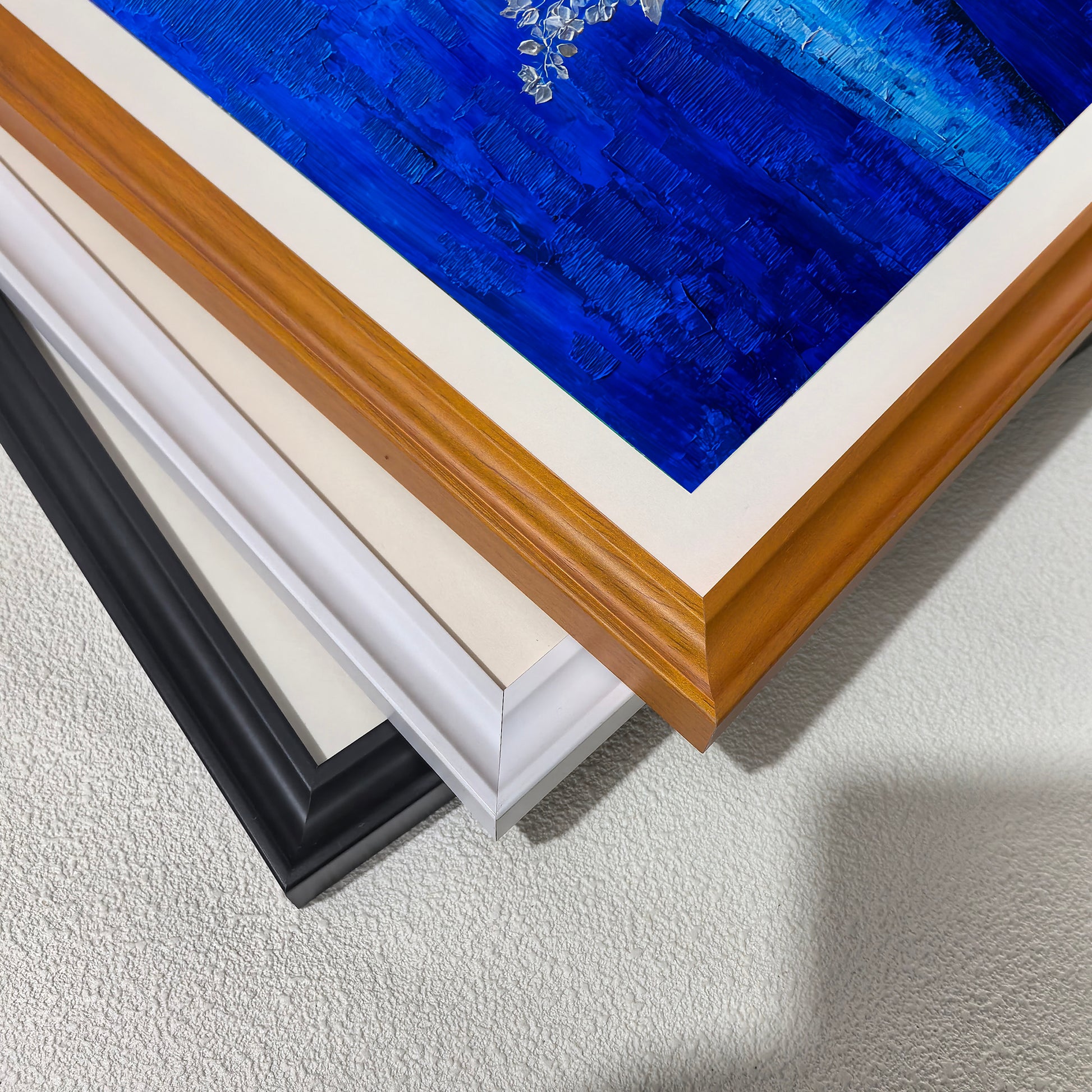 A close-up, stacked view of the corners of four three wooden picture frames: black, white, and light brown (wood grain), showing the frame profiles."
