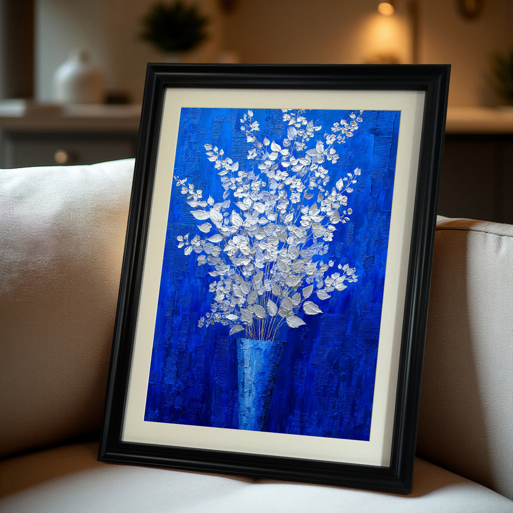A close-up, angled view of a vertical, black-framed painting of silver-white flowers on a royal blue background with a white mat, resting on a beige sofa.