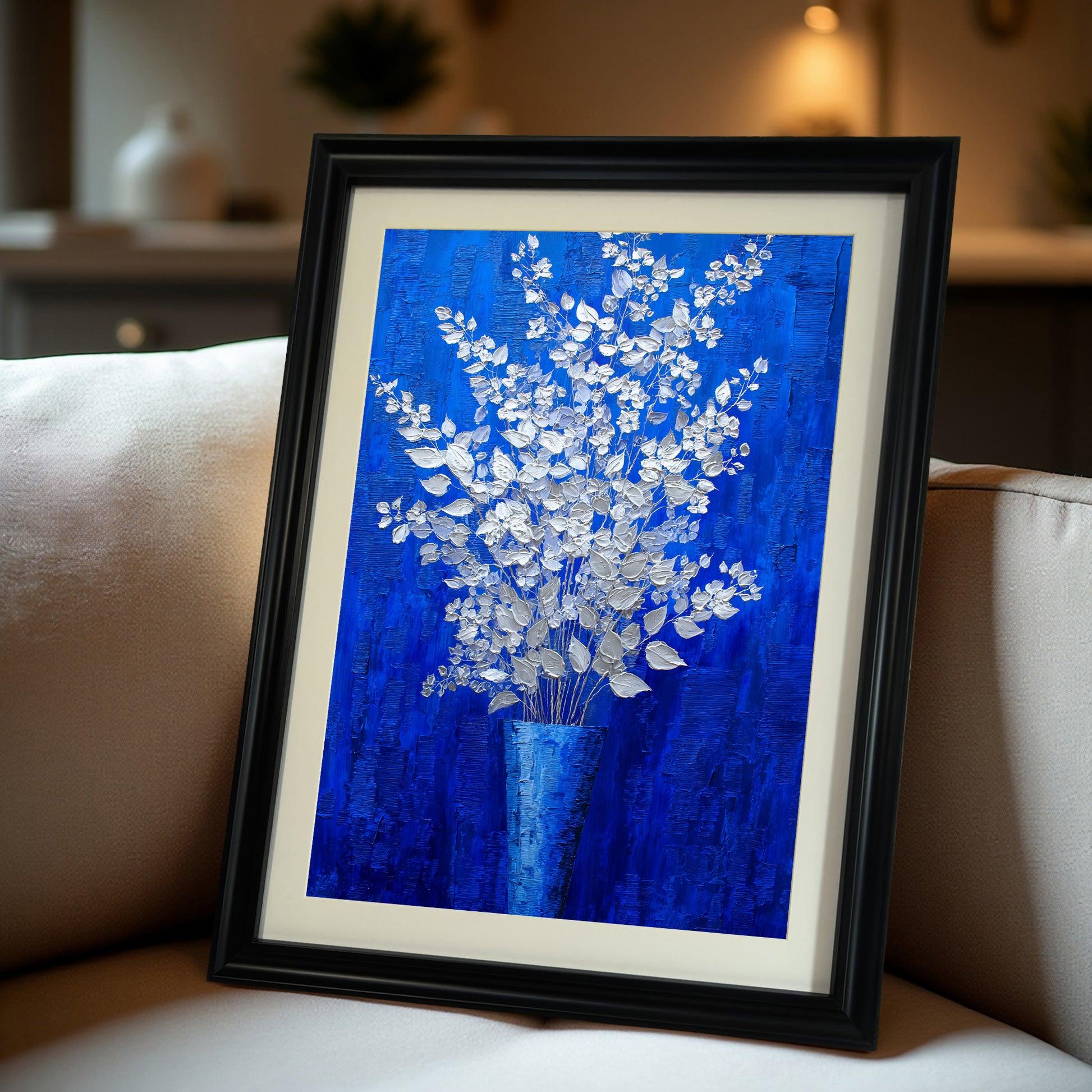 A close-up, angled view of a vertical, black-framed painting of silver-white flowers on a royal blue background with a white mat, resting on a beige sofa.