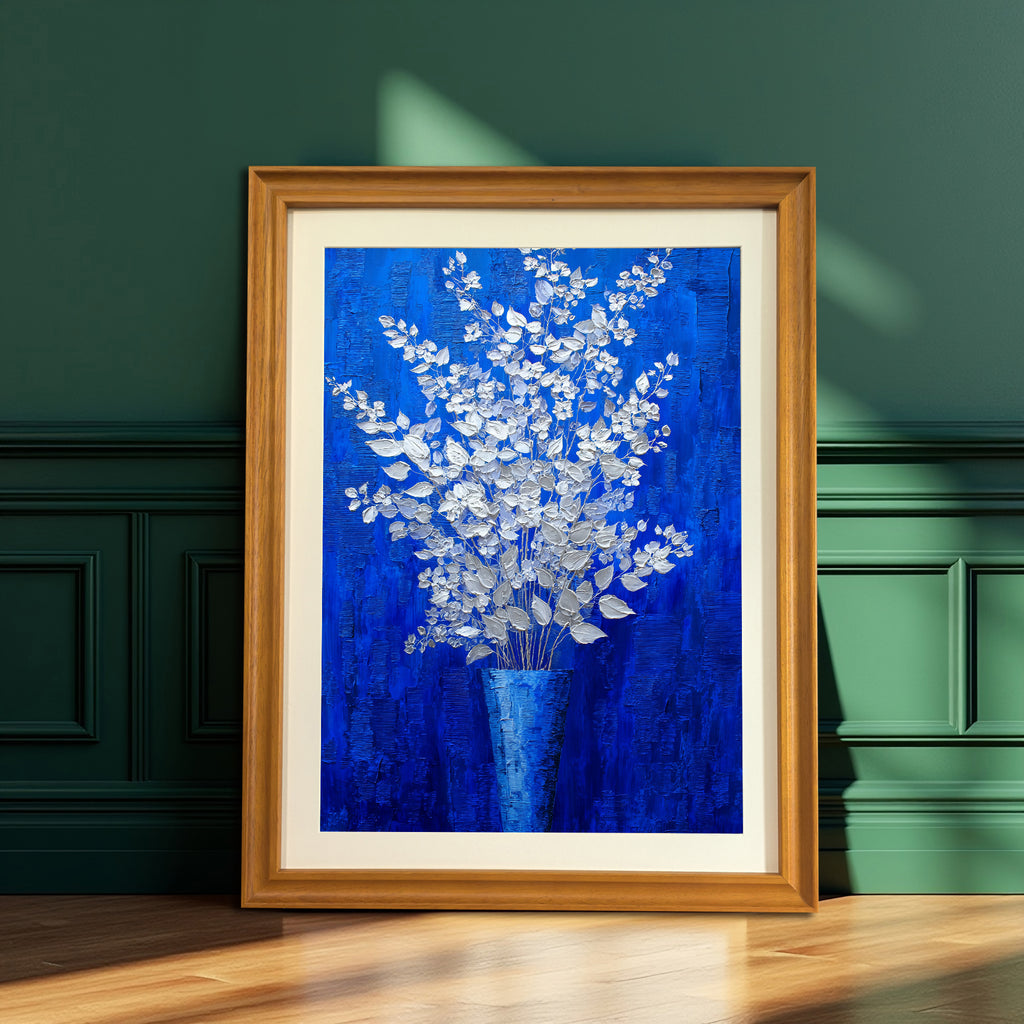 A vertical, framed oil painting of silver-white flowers in a blue vase against a royal blue background. The painting is in a light brown wood frame and is leaning on a hardwood floor against a dark green paneled wall with strong window light.