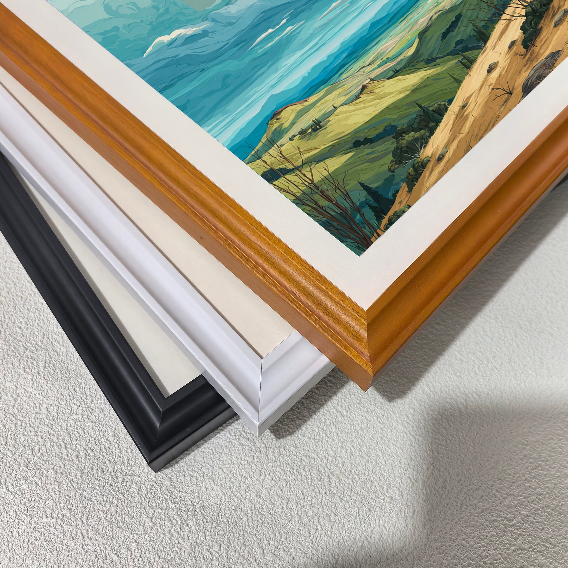Stacked close-up of different wood frame options for art prints: black, white, and natural light wood (oak/maple). Shows the frame molding detail and finish against the landscape art.