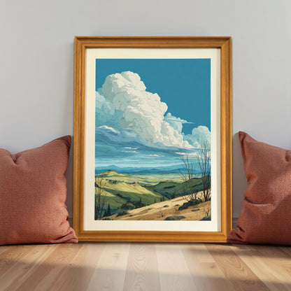 Rolling hills landscape art print in a natural light wood frame (oak/maple), leaning against a gray wall on the floor between two terracotta-colored pillows, demonstrating a cozy, warm decor style.