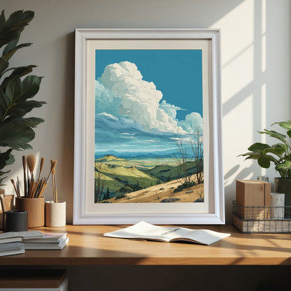 Rolling hills landscape art print in a white wood frame sitting on a wood desk next to art supplies and houseplants, showcasing a bright home office or studio setting.