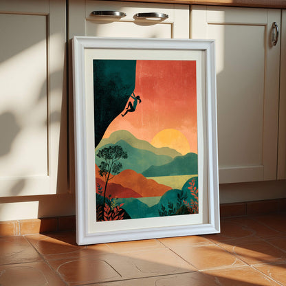Vertical wall art print of a female climber silhouette on a cliff against a red and orange sunset over layered green hills, framed in a thick white wooden frame, leaning against cream kitchen cabinets.