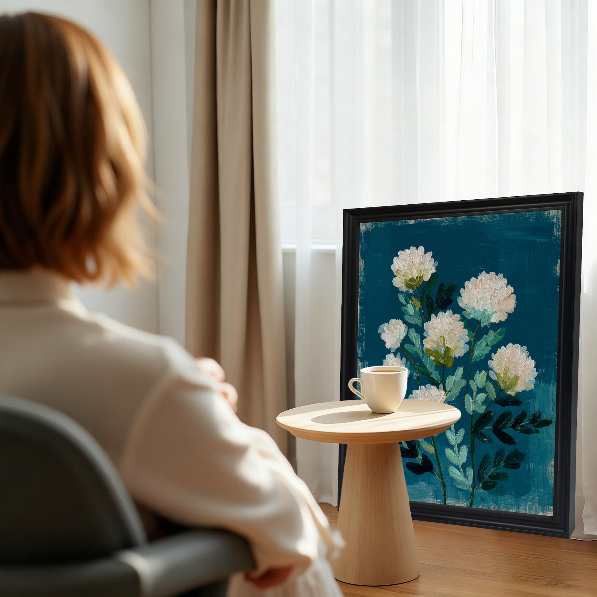 A black-framed art print leaning against a window with light curtains. The abstract white floral print is positioned next to a small wooden table with a coffee cup, with a woman looking towards it in the foreground.