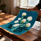 Close-up of hands holding an unframed, slightly curved art print of white abstract flowers and green leaves against a textured teal background, lying on a rustic wooden table.