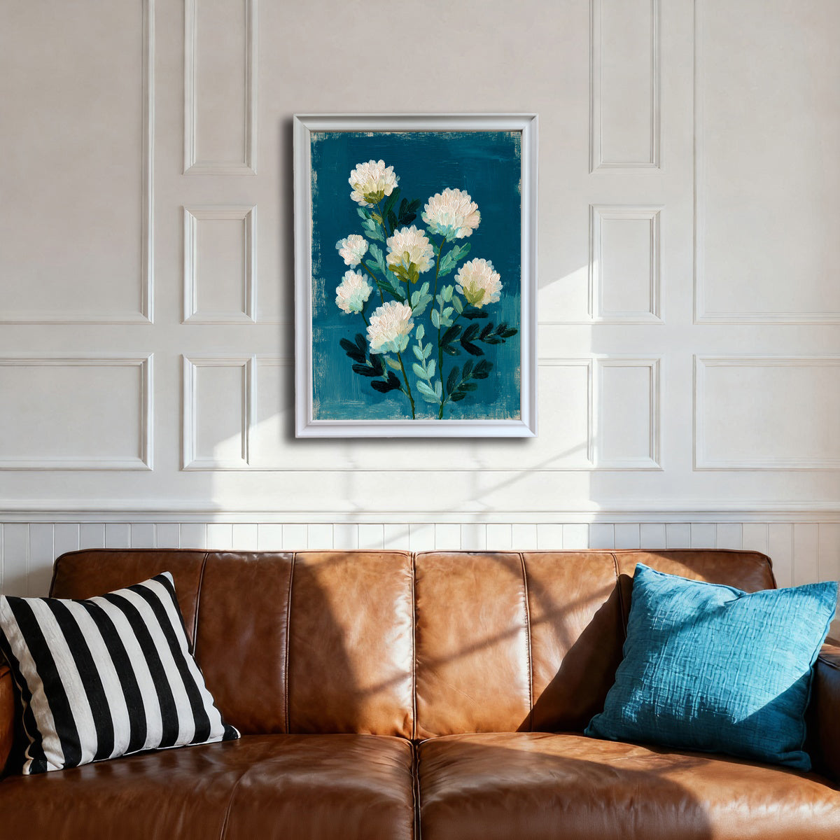 A white-framed art print hanging above a brown leather sofa. The white paneled wall provides a contrast to the deep teal-blue of the abstract floral artwork, which features white impasto flowers.