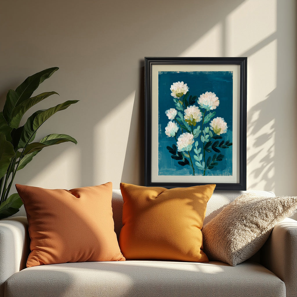 A black-framed art print resting on a white sofa with orange and gold pillows. The artwork is a vertical abstract floral painting featuring white, fluffy flowers on green stems against a vivid teal backdrop.