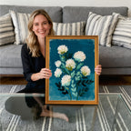A smiling woman sitting on the floor, holding a gold-framed art print. The painting features abstract white flowers with green leaves against a deep, textured teal-blue background.
