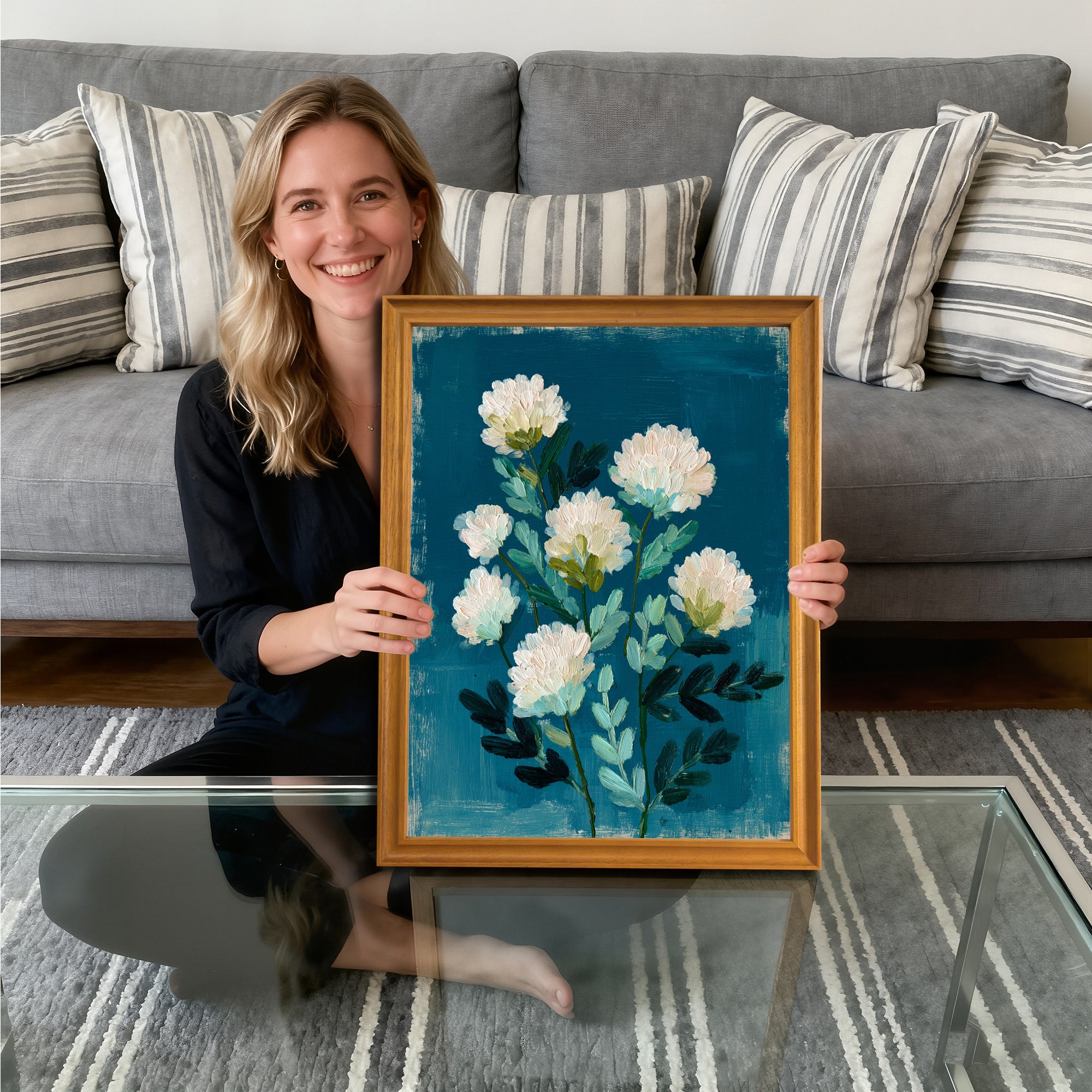 A smiling woman sitting on the floor, holding a gold-framed art print. The painting features abstract white flowers with green leaves against a deep, textured teal-blue background.