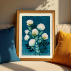 A wood-framed art print resting on a beige sofa between a dark blue pillow and a yellow pillow. The artwork shows an abstract floral pattern with white blooms and teal leaves against a deep blue canvas.