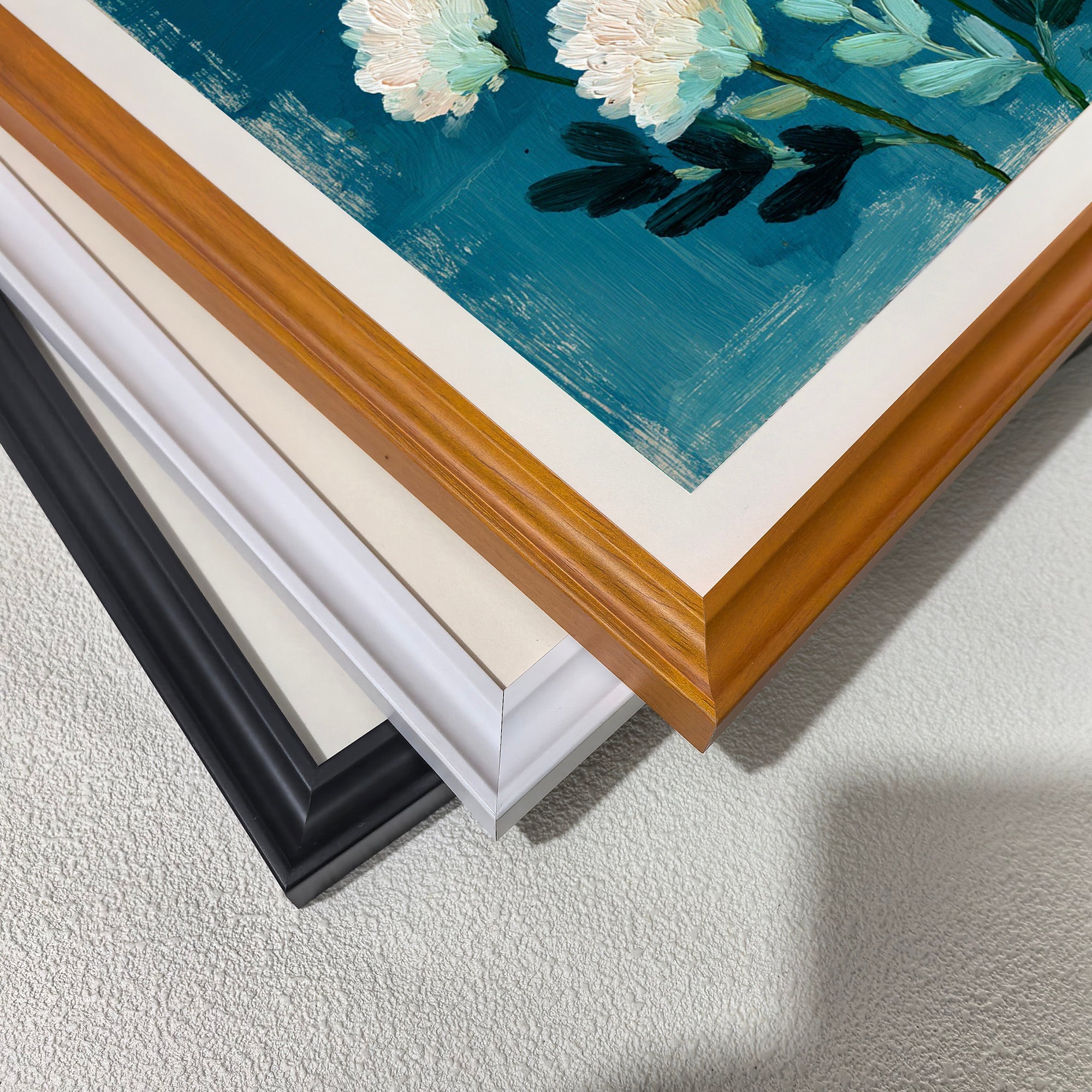 A stack of four frame corners: black, off-white, white, and a light wood grain (brown). This displays the available framing options for the abstract white floral art print.