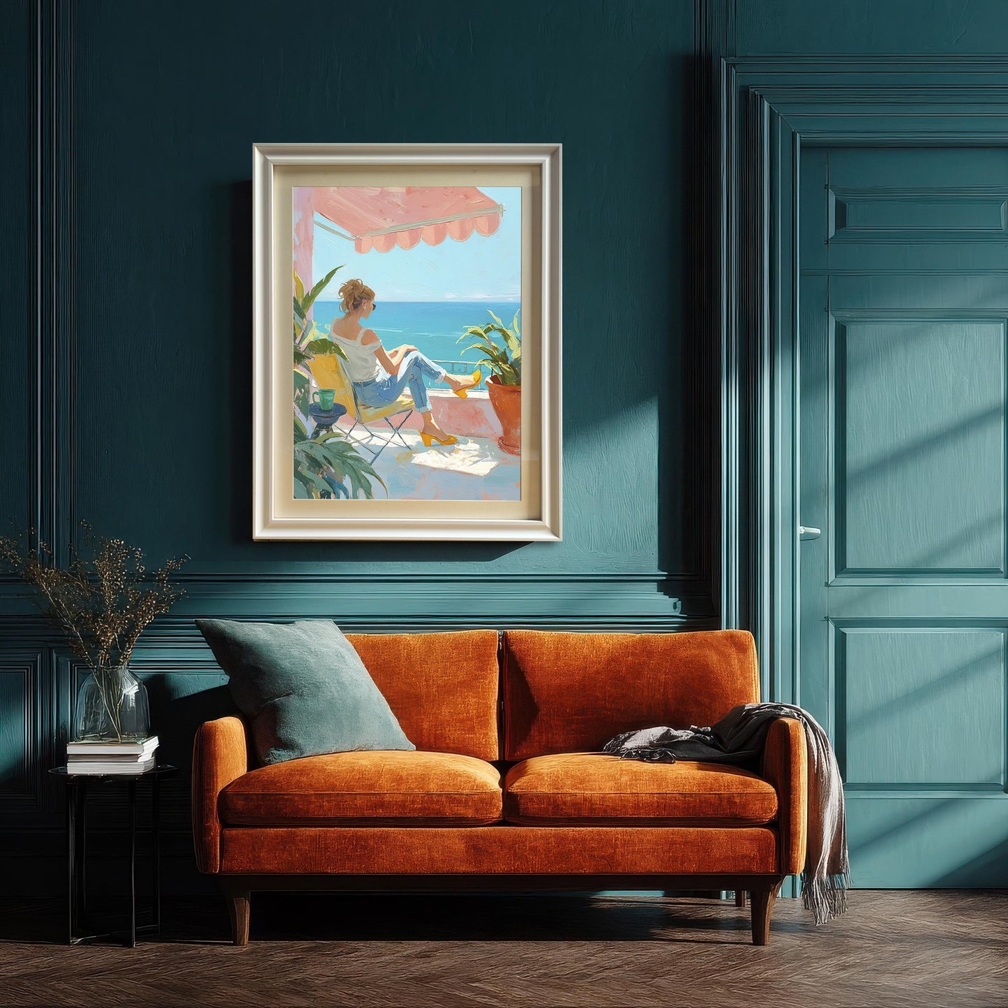 A large wall art print with a white frame hanging on a dark teal wall above an orange two-seater sofa. The painting shows a woman on a balcony looking at the sea. A small side table with a vase of dried flowers is to the left of the sofa.