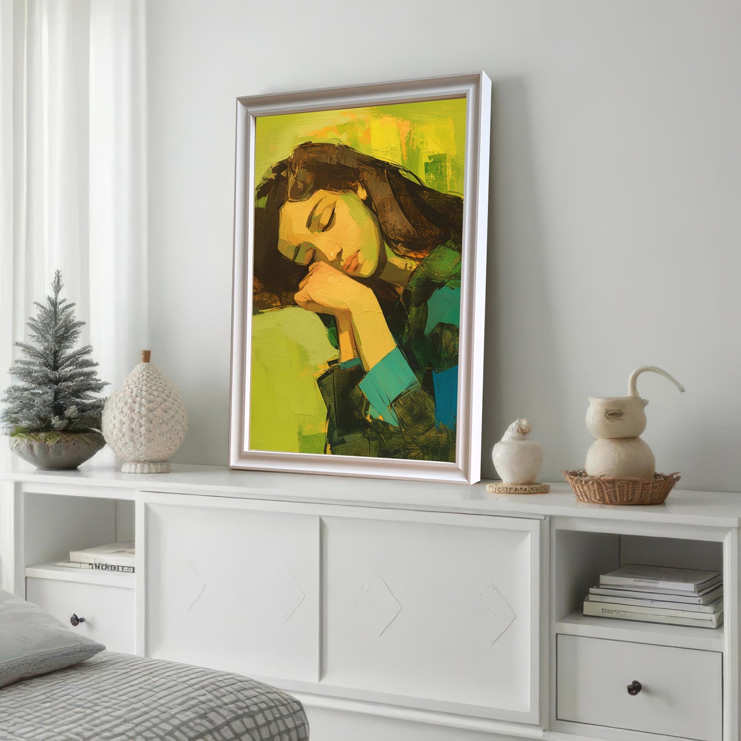 A framed wall art print with a white frame is leaning against a white wall on a white cabinet. The painting is an abstract portrait of a sleeping woman. The cabinet is decorated with various white and gray decorative objects.