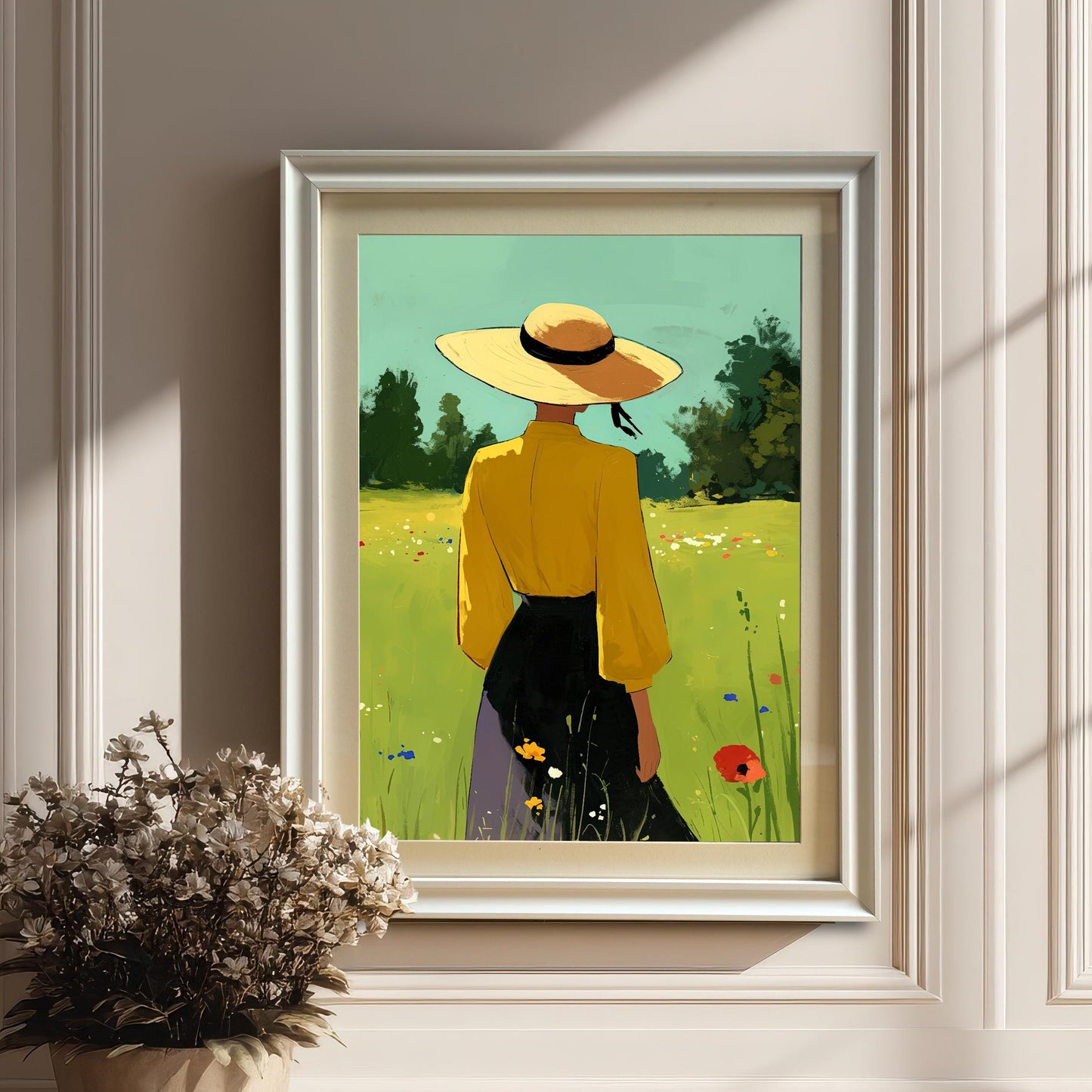 A white-framed art print with a white mount, depicting a person in a yellow shirt and straw hat in a field of flowers, hanging on a white wall above a potted plant with white flowers. The image is set in a bright room with architectural details on the wall.