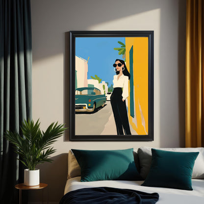 A framed piece of wall art hangs above a bed. The art depicts a stylish woman in a white shirt and black pants standing on a street next to a vintage teal car. The background is a bright yellow wall and a clear blue sky. The frame is black.
