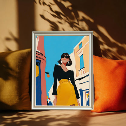 A framed art print with a white frame and no mat rests on a sofa between two plush pillows, one mustard yellow and one orange. The art shows a stylish woman in a black shirt and yellow skirt standing on a street.