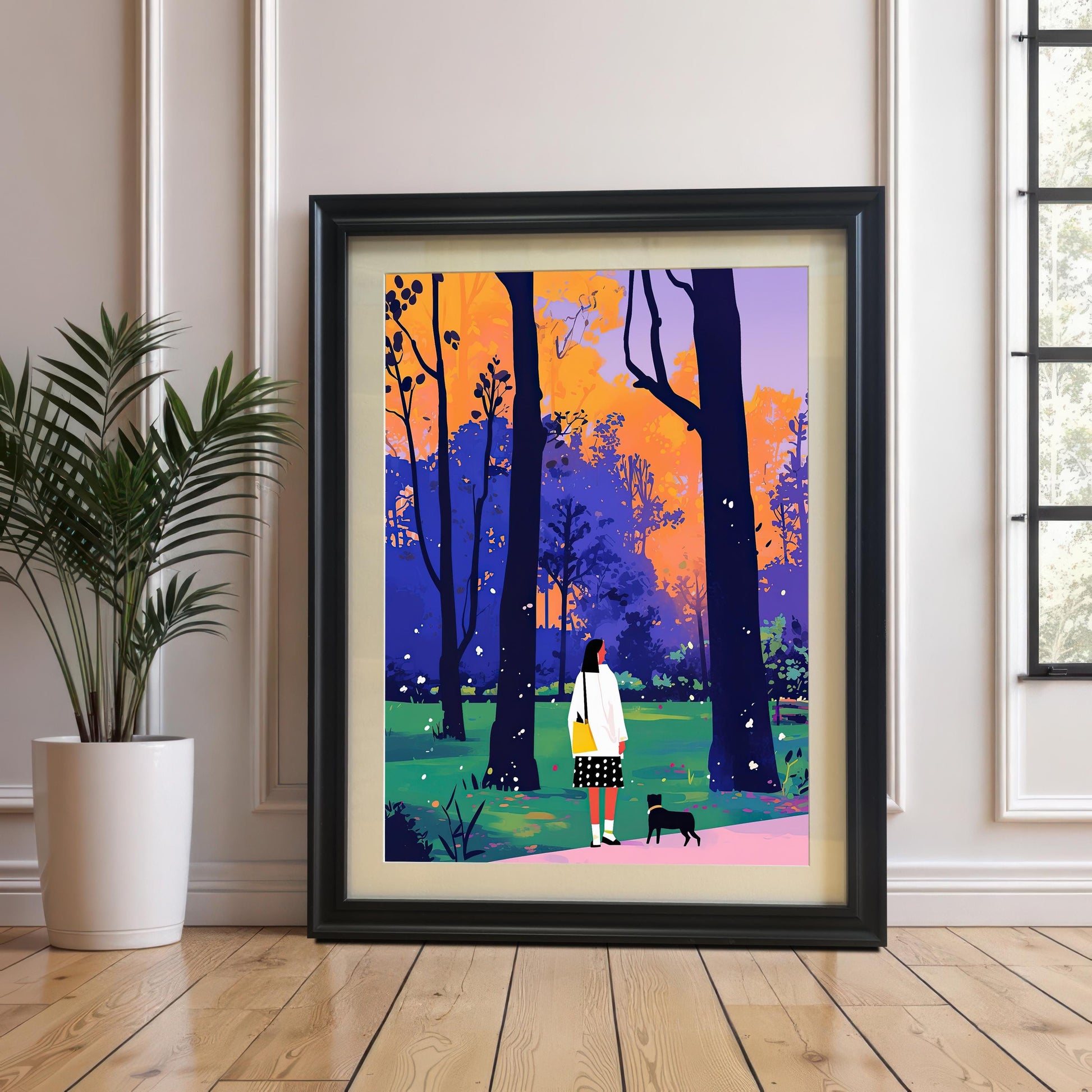 A framed art print with a black frame and a white mat leans against a white paneled wall and rests on a light-colored wooden floor. The art depicts a woman and a small black dog walking through a park with colorful trees. A potted plant sits on the floor to the left of the frame.