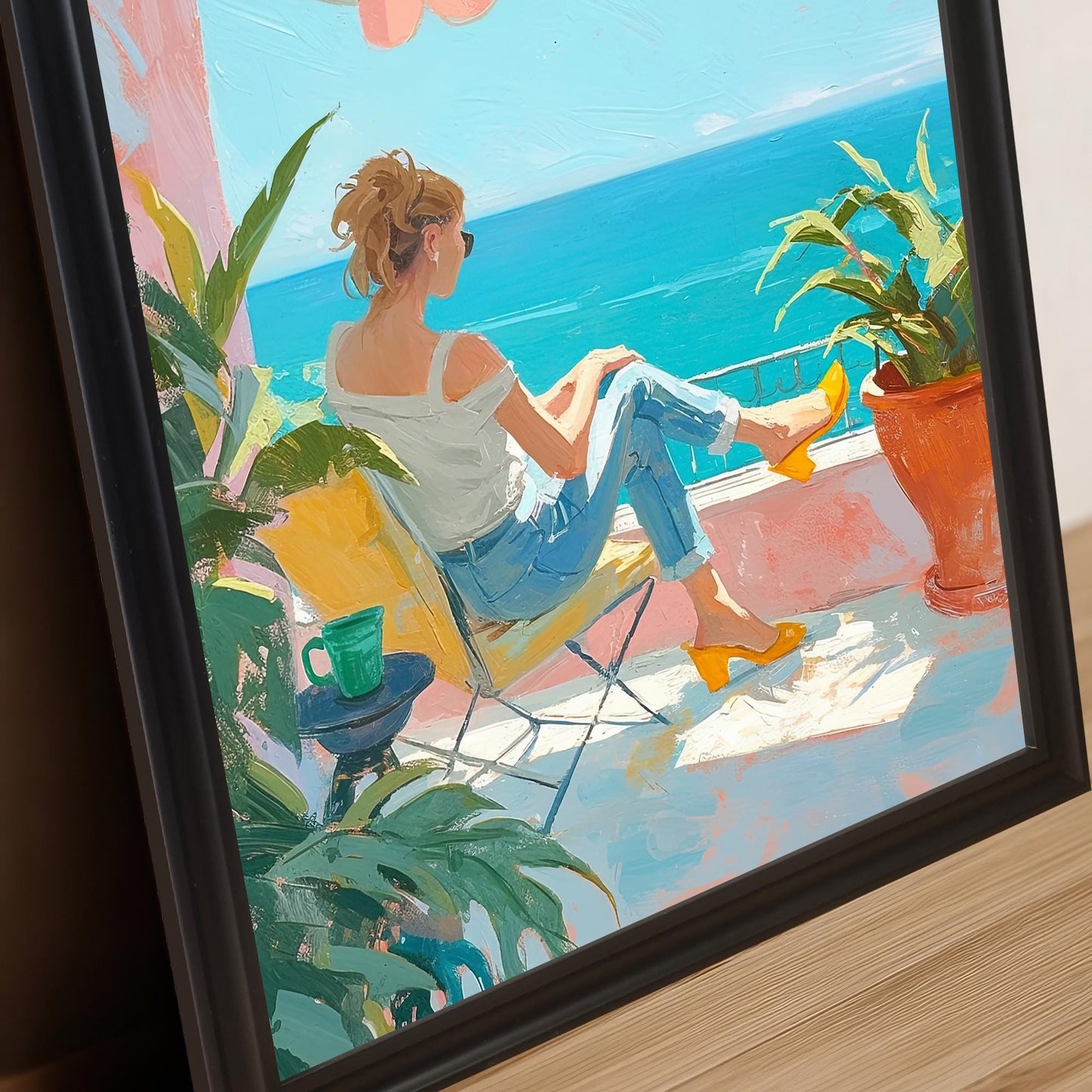 A close-up image of a wall art print with a black frame. The print is of a woman on a balcony looking at the ocean. The frame is positioned at an angle, resting on a wooden surface, showing the detail of the artwork and the frame's edge.