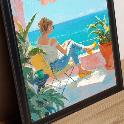 A close-up image of a wall art print with a black frame. The print is of a woman on a balcony looking at the ocean. The frame is positioned at an angle, resting on a wooden surface, showing the detail of the artwork and the frame's edge.