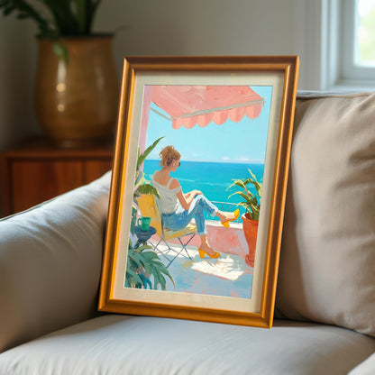 A close-up shot of a single wall art print with an orange wood frame, leaning against a white sofa cushion. The painting depicts a woman in a white shirt and jeans, seated on a balcony chair, looking out at the ocean.