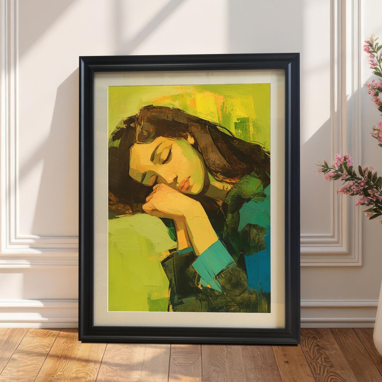 A vertical wall art print with a black frame, resting on a light wood floor against a white wall. The painting is an abstract portrait of a woman with dark hair, sleeping with her head on her hands, against a bright yellow and green background. A tall plant with pink flowers in a white vase is visible to the right.