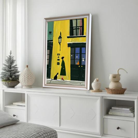 A framed wall art print featuring a woman in a green coat and a black cat walking on a snowy street in front of a bright yellow building. The print is framed in white and is displayed on a white dresser.