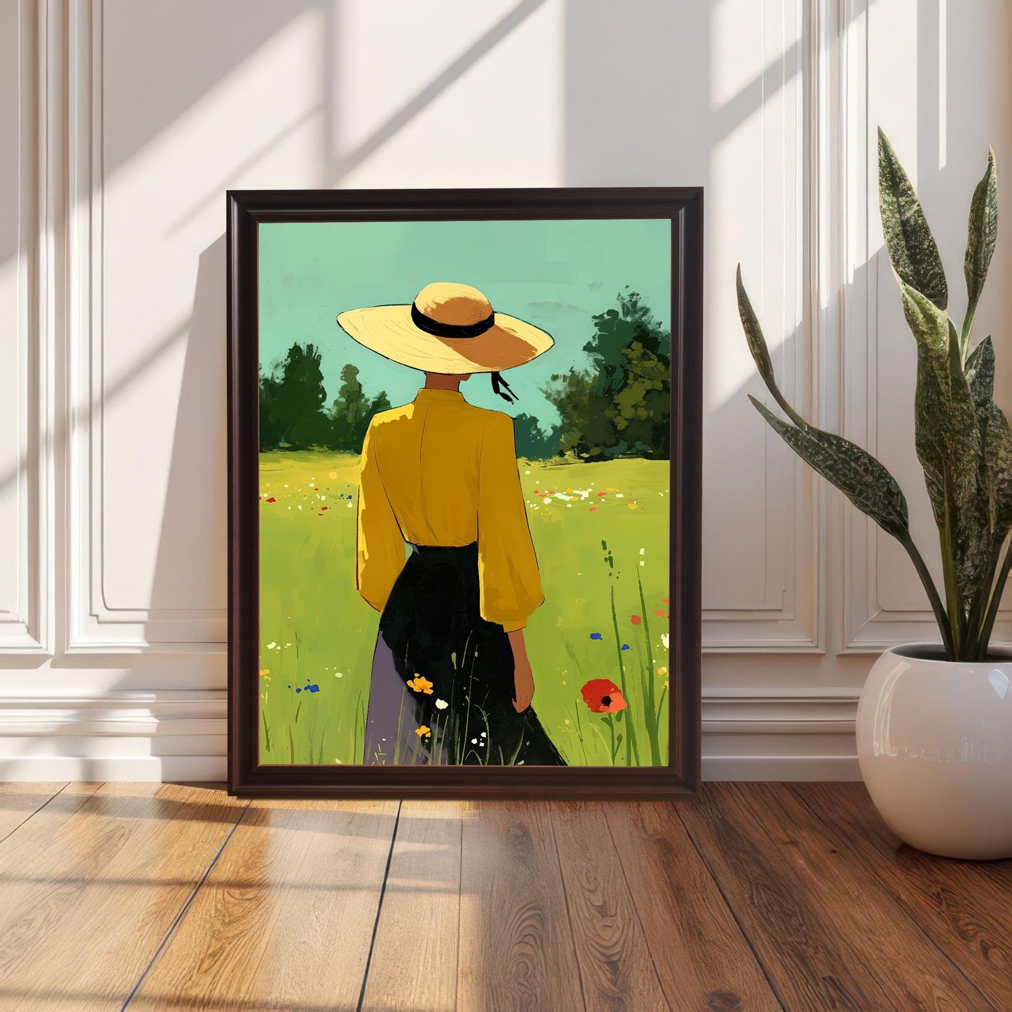 A single art print with a dark coffee-colored wood frame leaning against a white wainscoting wall on a hardwood floor. The print shows a person in a yellow shirt and a wide-brimmed hat standing in a field of green grass and colorful flowers. A large green potted plant is visible to the right of the frame.