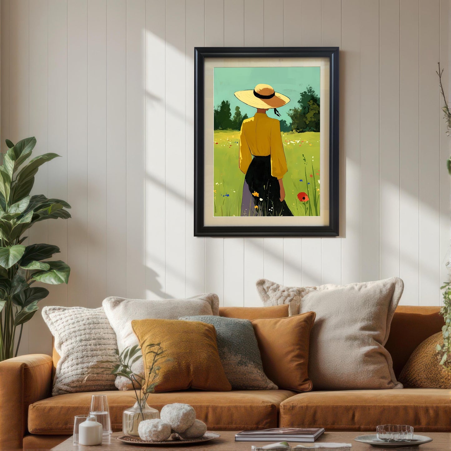 A black-framed and mounted art print of a person in a yellow shirt and straw hat in a field of flowers, hanging on a white wall above a brown couch with multiple pillows. Two large potted plants are visible on either side of the couch. The setting is a cozy living room.