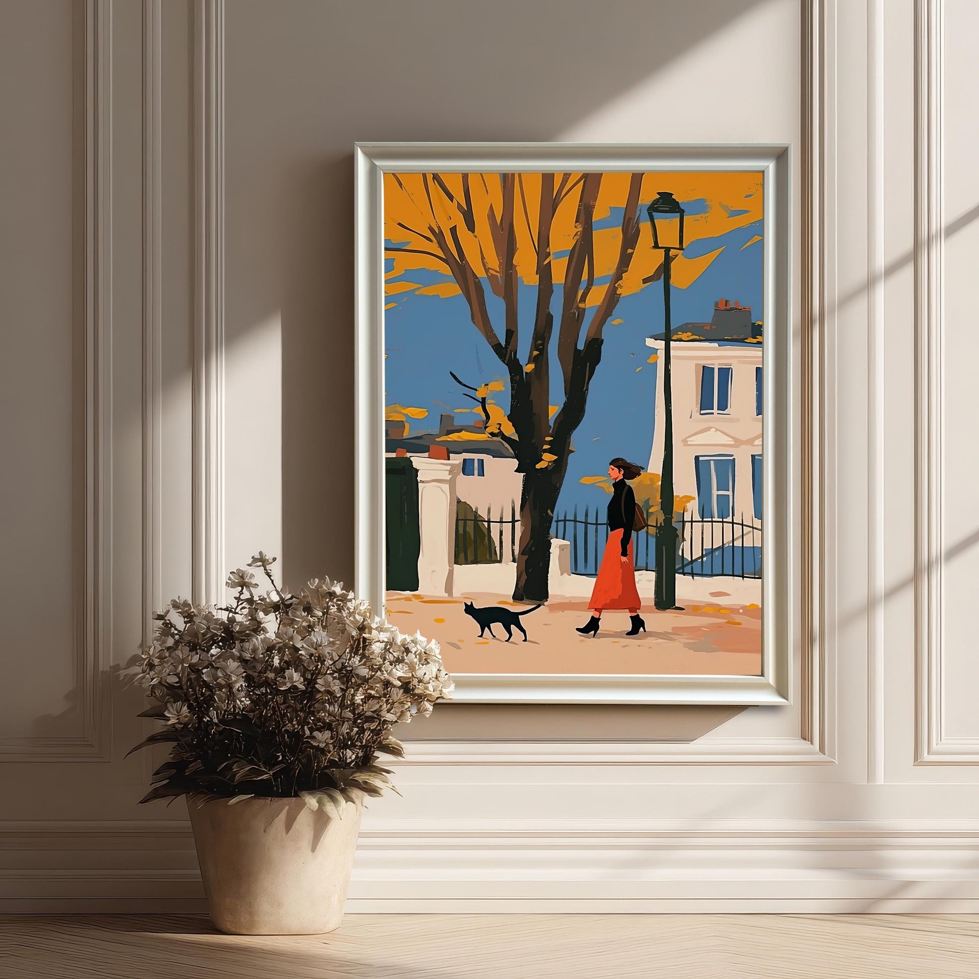 A framed art print of a woman and a black cat walking on an autumn street, hanging on a white paneled wall next to a potted plant.