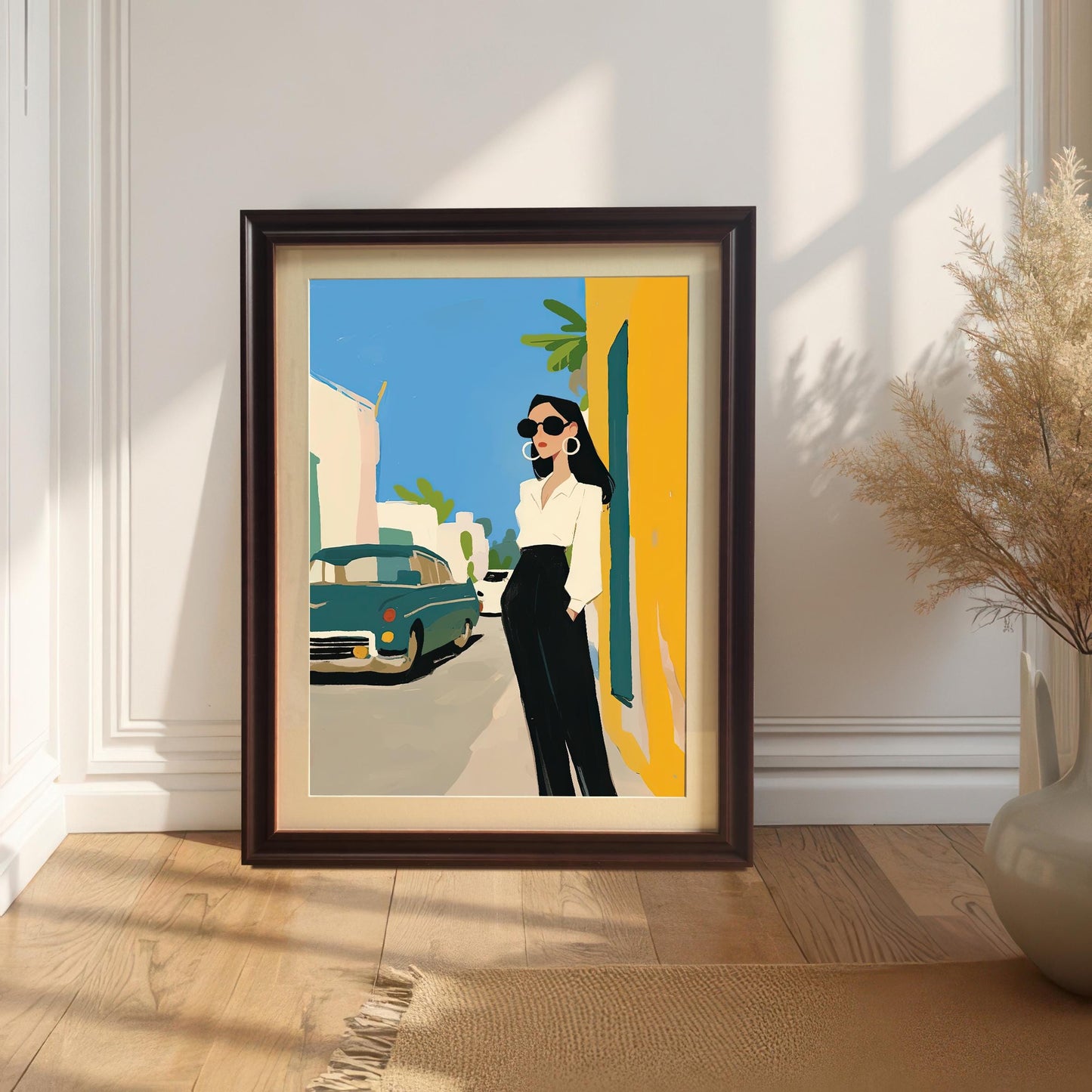 A framed art print with a coffee brown frame and a white mat leans against a white wall and rests on a light-colored rug. The art shows a stylish woman in a white shirt and black pants standing on a street next to a vintage teal car. A large vase with dried grasses sits next to the frame.