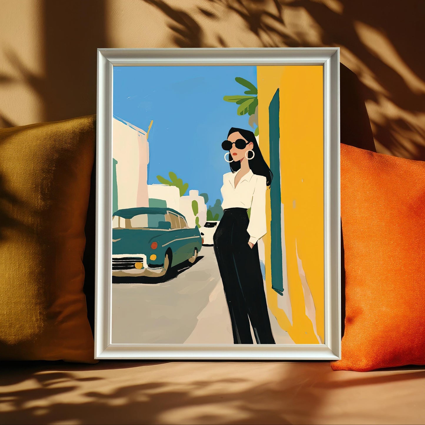 A framed art print with a white frame and no mat rests on a sofa between two plush pillows, one mustard yellow and one orange. The art shows a stylish woman in a white shirt and black pants standing on a street next to a vintage teal car.