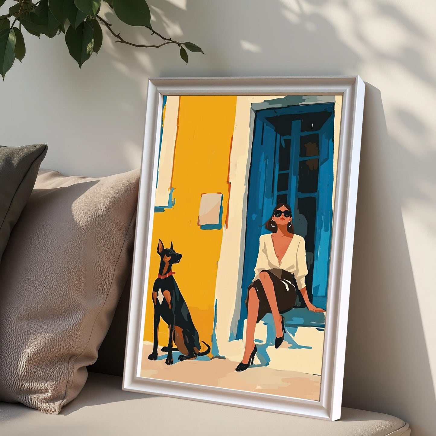 A framed art print with a white frame and no mat rests on a sofa between two plush pillows, one gray and one beige. The art shows a woman in a white shirt and black skirt sitting on a step next to a Doberman Pinscher dog.