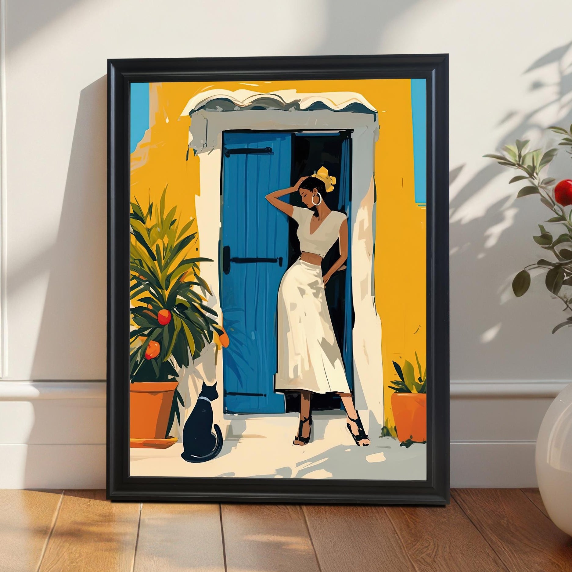 A vertical framed artwork in a black frame leans against a wall on a wooden floor. The artwork depicts a woman in a white outfit, standing in a doorway with a bright blue door and a yellow building. A black cat sits nearby. A potted plant with red berries is next to the frame.