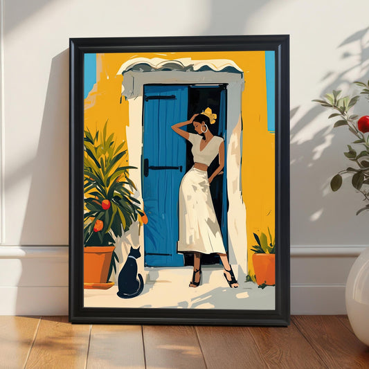 A vertical framed artwork in a black frame leans against a wall on a wooden floor. The artwork depicts a woman in a white outfit, standing in a doorway with a bright blue door and a yellow building. A black cat sits nearby. A potted plant with red berries is next to the frame.