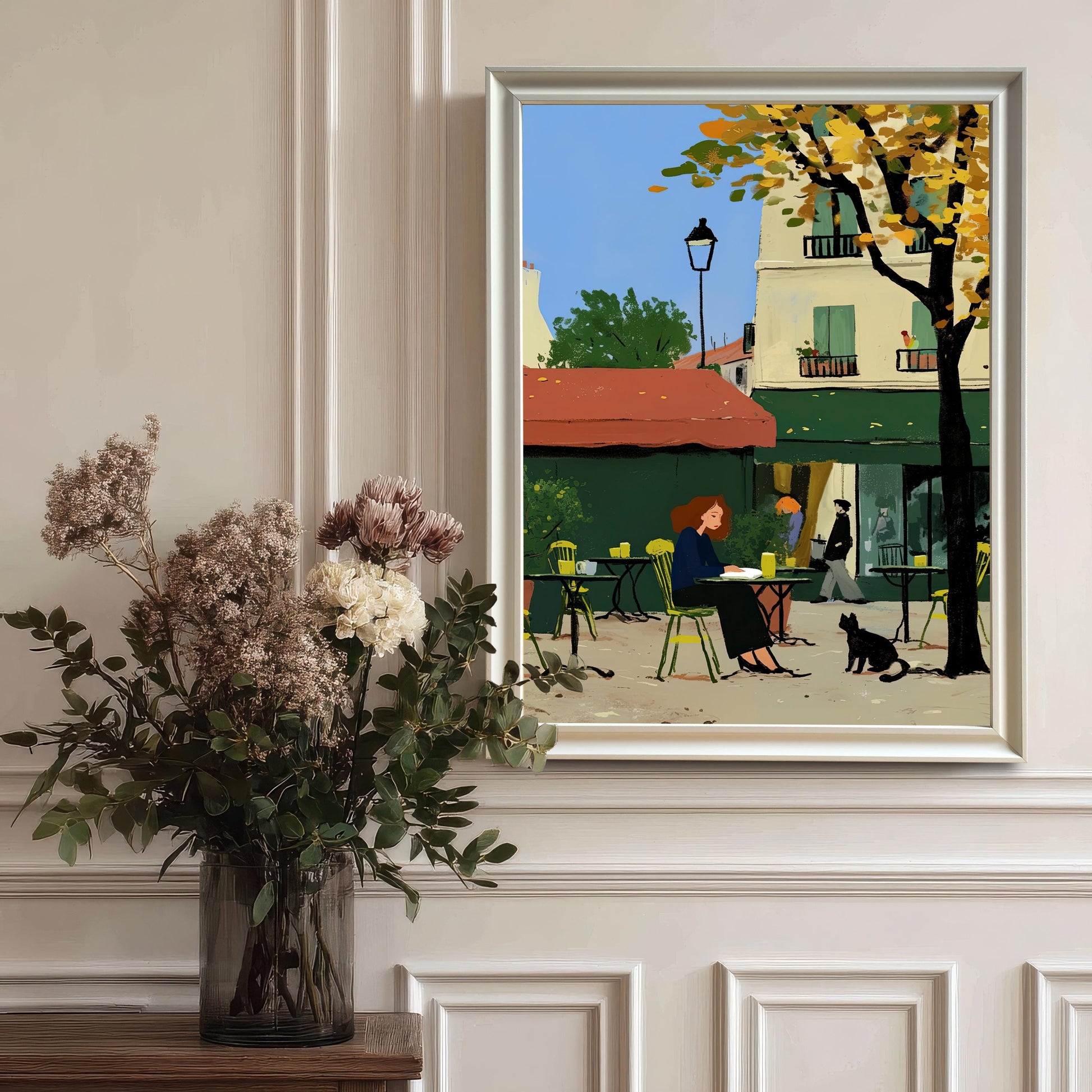 A framed artwork in a white frame hangs on a white paneled wall. The artwork depicts a woman sitting at an outdoor cafe table with a black cat nearby. The background features a building with a red awning and a tree with yellow autumn leaves. A large vase with dried flowers is in the foreground on the left.