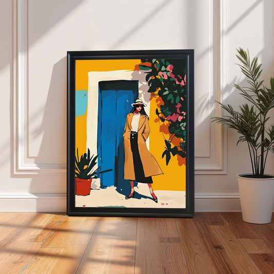 A framed art print with a black frame and a white mount is leaning against a white paneled wall on a wooden floor. The art depicts a woman in a tan coat standing in front of a yellow building with a blue door. A green potted plant is visible on the right.